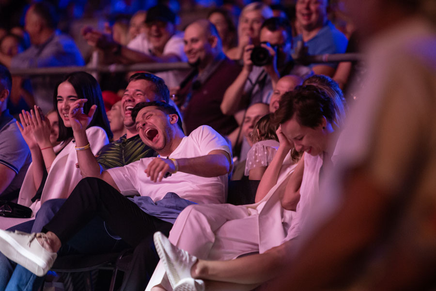 Crowd laughing enthusiastically at a comedy show, with people in the front row leaning back in laughter under stage lighting.