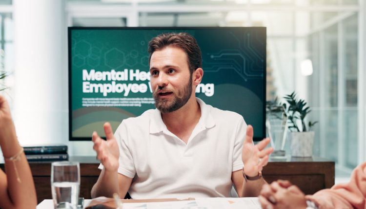 Man speaking during a meeting about mental health and employee well-being, with a presentation screen behind him displaying the topic.