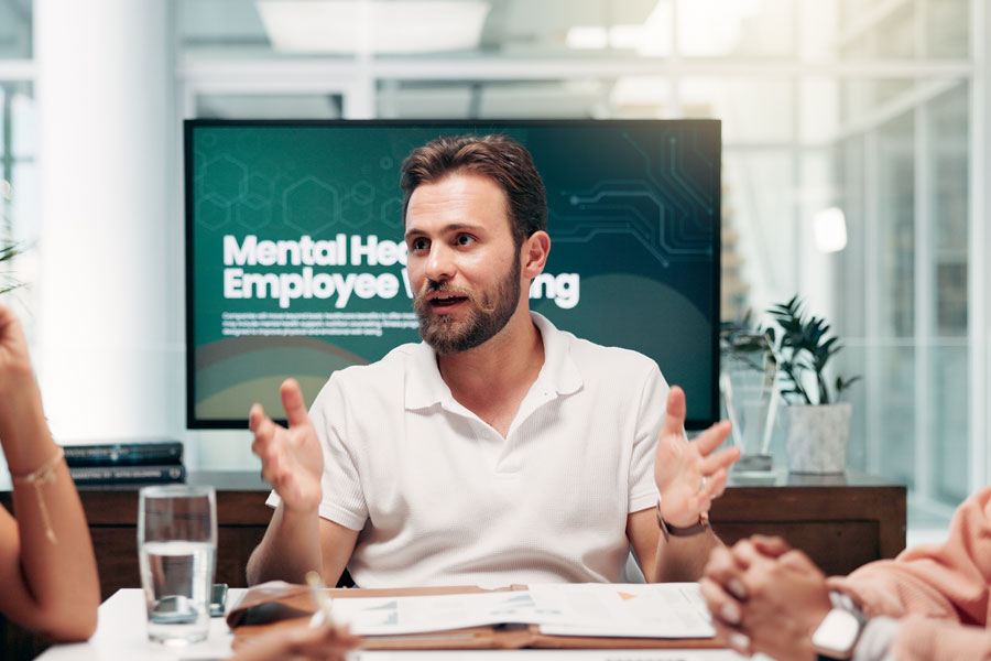 Man speaking during a meeting about mental health and employee well-being, with a presentation screen behind him displaying the topic.