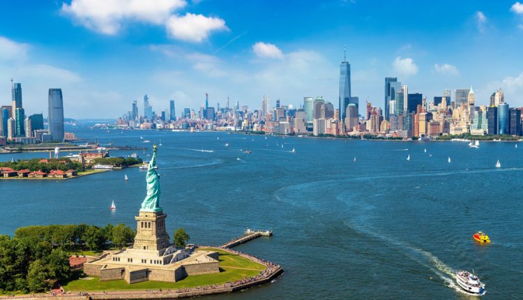 Aerial view of the Statue of Liberty on Liberty Island with Fort Wood's star-shaped base, Upper New York Bay, and the Manhattan skyline in the distance under clear blue skies. Sailboats and tourist ferries are visible on the harbor waters.