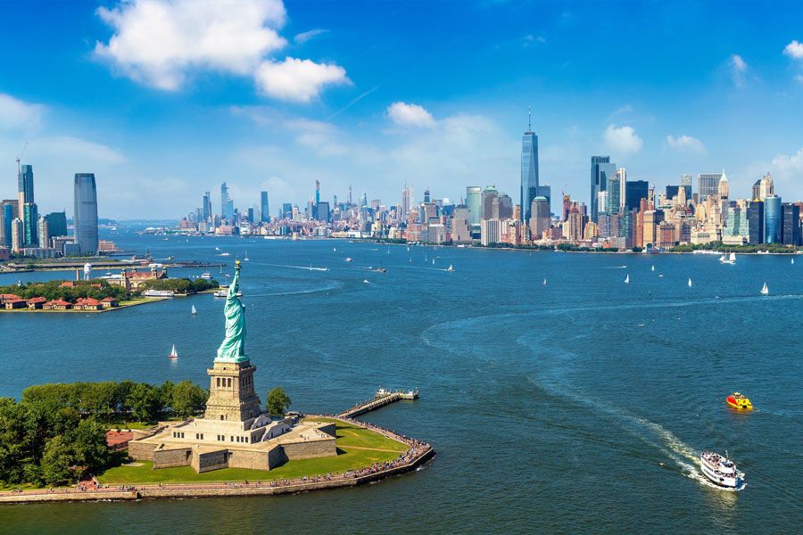 Aerial view of the Statue of Liberty on Liberty Island with Fort Wood's star-shaped base, Upper New York Bay, and the Manhattan skyline in the distance under clear blue skies. Sailboats and tourist ferries are visible on the harbor waters.