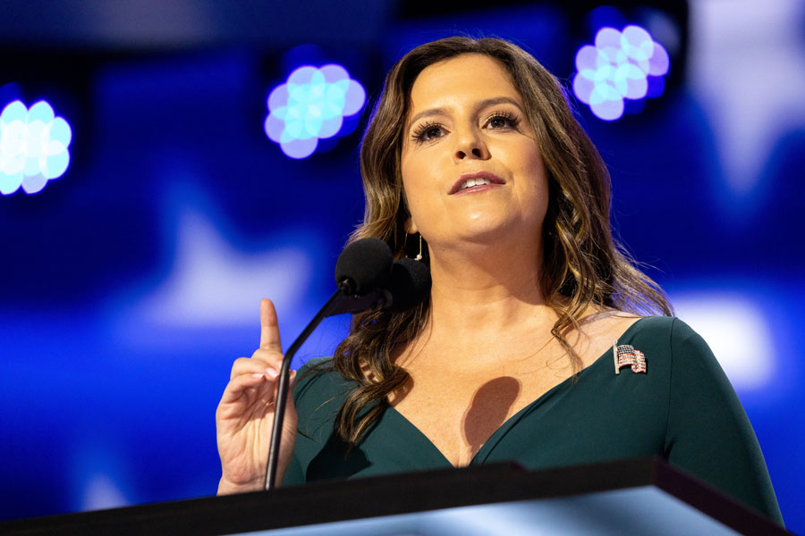 Elise Stefanik speaking at a podium under bright stage lights, wearing a dark green dress and an American flag pin.