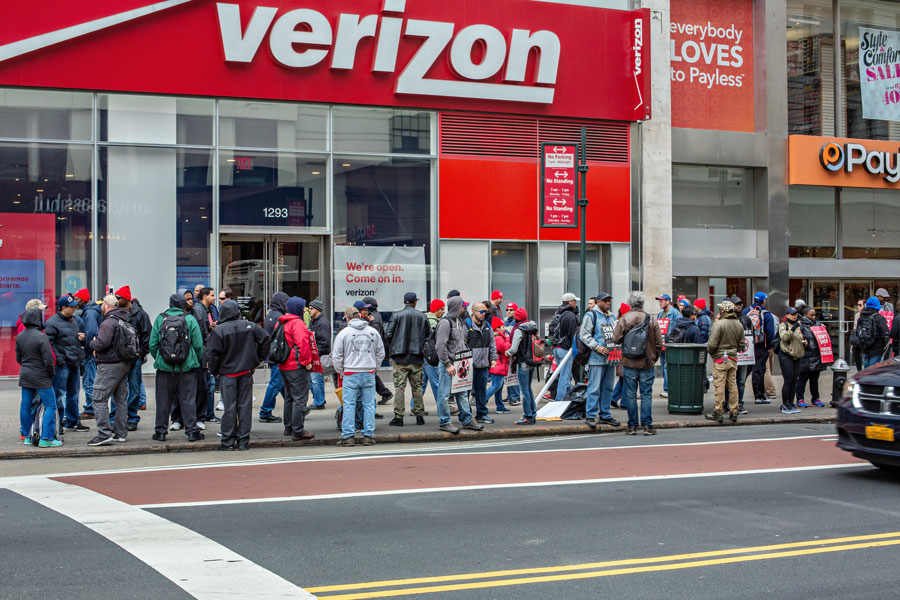 A large group of Verizon workers and union supporters stand outside a Verizon store in New York City during a 2016 strike, holding signs and gathering along the sidewalk.