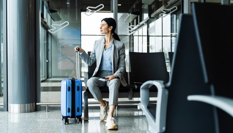 Business woman sitting in airport and waiting for starting her travel voyage. Woman with luggage is holding handle of the suitcase and looking around airport terminal