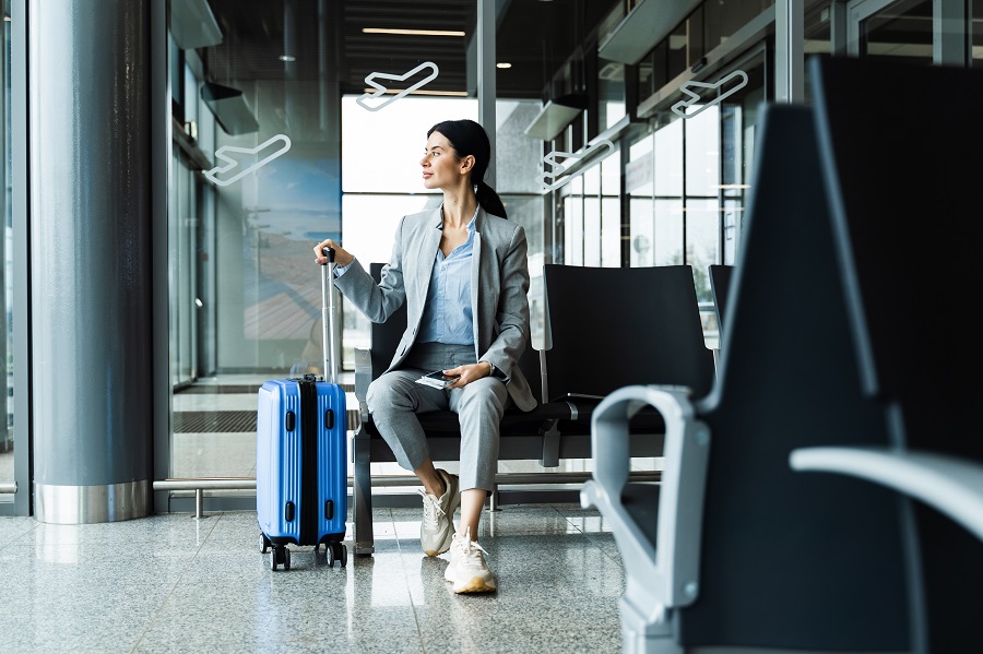 Business woman sitting in airport and waiting for starting her travel voyage. Woman with luggage is holding handle of the suitcase and looking around airport terminal