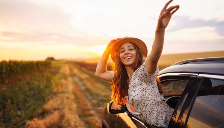 Young woman smiling and waving from a car window while traveling on a scenic road at sunset, symbolizing leisure travel and tourism.