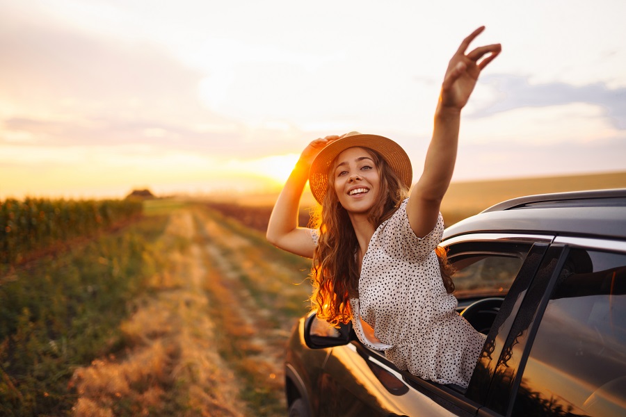 Young woman smiling and waving from a car window while traveling on a scenic road at sunset, symbolizing leisure travel and tourism.