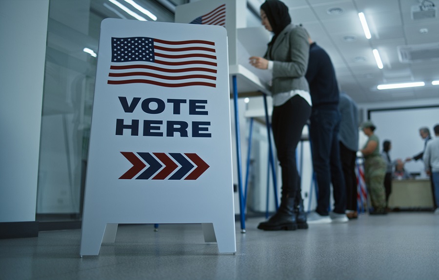 Voters casting ballots at a polling location in the United States with a Vote Here sign in the foreground.