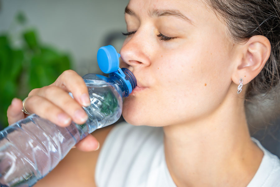 Woman drinking from a plastic bottled water at home, illustrating rising bottled-water use and growing concerns about microplastics and chemical exposure.