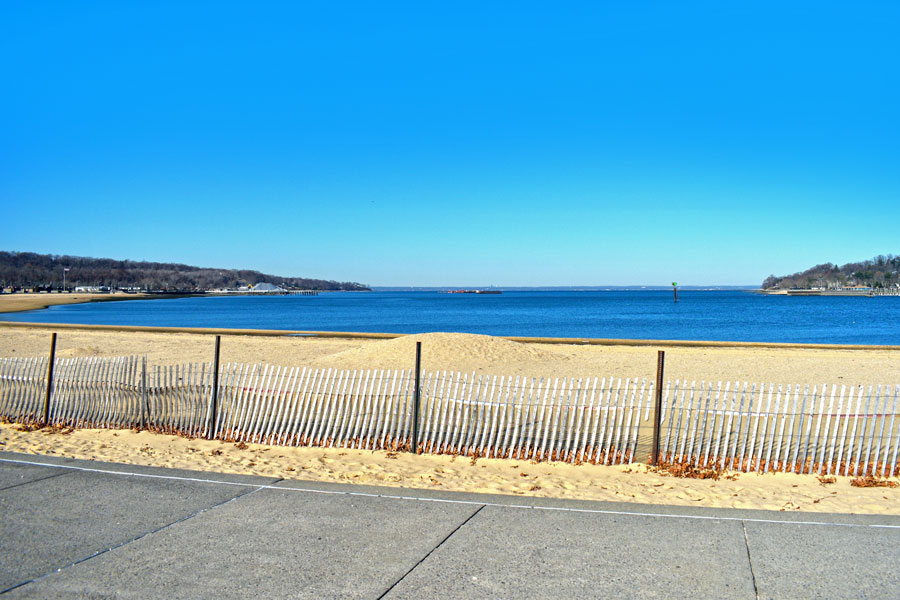 View of North Hempstead Beach Park with sandy shoreline, fencing, and Hempstead Harbor in the background.