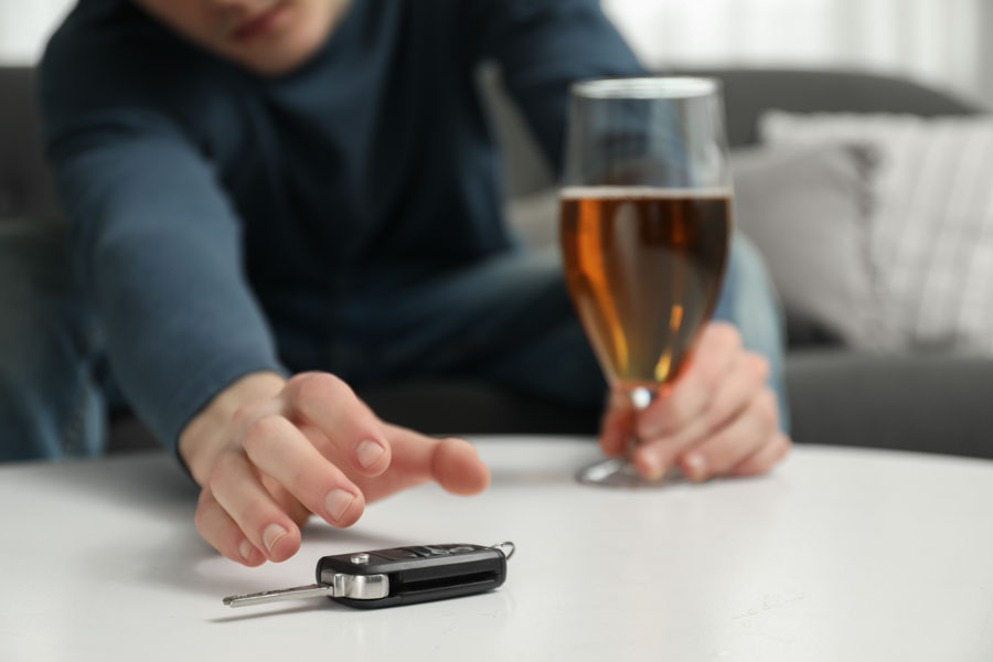 Man holding a glass of alcohol reaching toward car keys on a table, symbolizing the dangers of driving while intoxicated.