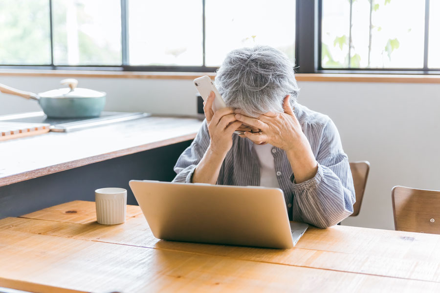 Elderly woman holding a phone and covering her face while seated at a laptop, representing emotional distress caused by a phone scam targeting seniors.