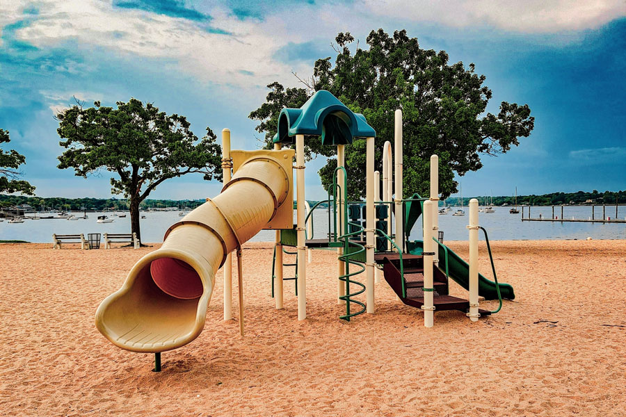 Playground structure on the sand at Manorhaven Beach Park with trees, boats, and Manhasset Bay in the background.