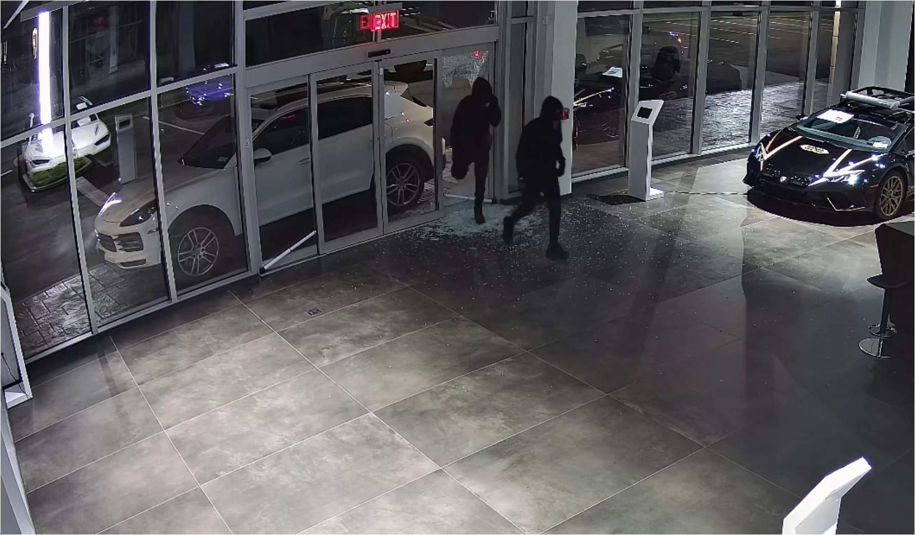 Surveillance image from police-released video shows two masked individuals inside an auto dealership showroom during an alleged overnight burglary in July 2025. Shattered glass is visible near the entrance doors as the suspects move through the showroom, where high-end vehicles are displayed under interior lighting.