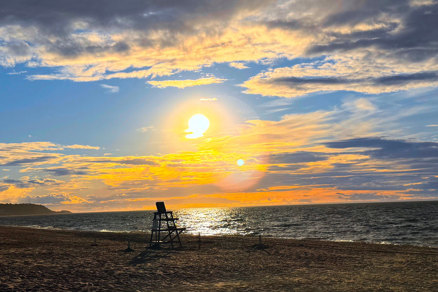 Sunset over Cedar Beach in Mount Sinai, New York, with a lone lifeguard chair overlooking the Long Island Sound as waves gently roll onto the shoreline. The sky glows with vibrant shades of orange, gold, and blue, creating a peaceful coastal scene along Long Island’s North Shore.