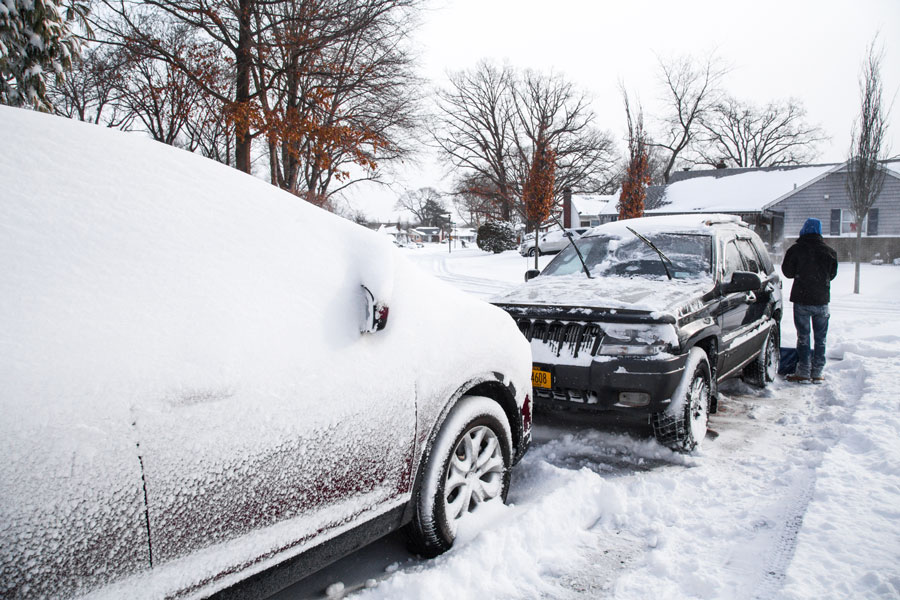A snow-covered residential street in East Williston, New York, after a winter storm, as a resident clears snow from parked vehicles in this quiet Nassau County neighborhood.