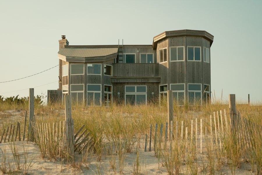 A modern beach house rises behind protected dunes in Dunewood on Fire Island, where sandy pathways, native beach grass, and quiet oceanfront living reflect the relaxed character of this secluded coastal community.