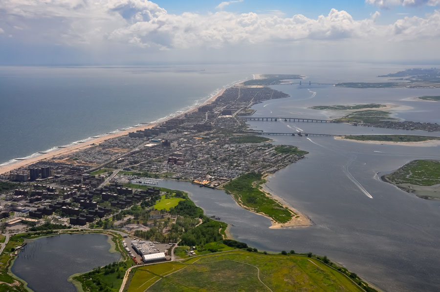 An aerial view looking west across the Rockaway Peninsula in Queens, New York, showing the Atlantic Ocean shoreline, Jamaica Bay, and the coastal communities of Rockaway Park, Belle Harbor, and Seaside. The narrow barrier peninsula highlights the unique geography that connects New York City’s waterfront neighborhoods with the greater Long Island South Shore region.
