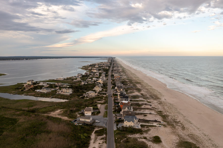 An aerial view looking east along Dune Road showcases the unique geography of Cupsogue Beach County Park, where the Atlantic Ocean borders one side of the barrier island and Moriches Bay stretches along the other. The park is one of Suffolk County's premier oceanfront destinations, known for its expansive sandy beach, camping, surf fishing, and protected coastal dunes.