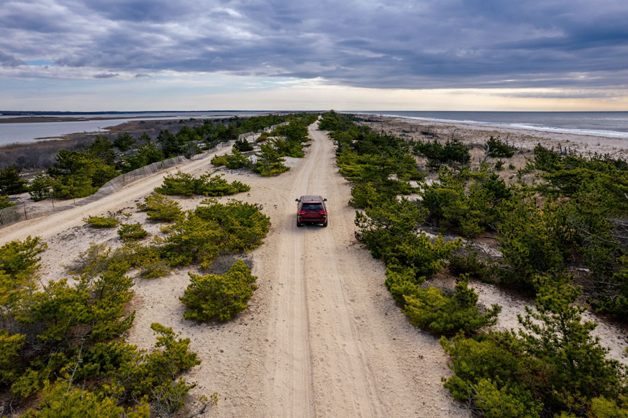 Smith Point County Park, NY, USA - April 10, 2022 - Aerial view of a red SUV driving along a sandy coastal road beside the ocean on Long Island, New York.