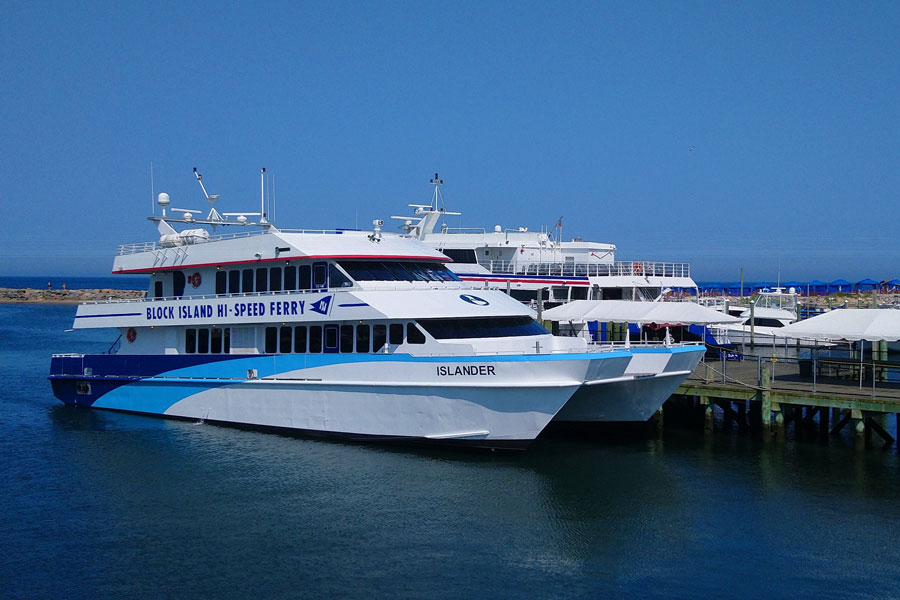 The Block Island Hi-Speed Ferry is docked at Block Island's waterfront terminal on a clear summer day, awaiting passengers for service between the island and mainland ports. The modern high-speed catamaran, along with additional ferry vessels moored nearby, highlights the important role ferry transportation plays in connecting Block Island with Rhode Island, Long Island, and other destinations throughout southern New England.