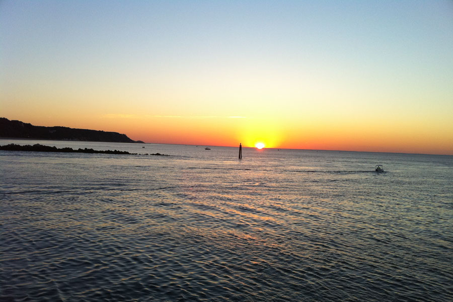 The sun sets over Long Island Sound at Cedar Beach–Mount Sinai, casting warm shades of gold and orange across the gently rippling water. Boats near the entrance to Mount Sinai Harbor add to the tranquil North Shore scene.