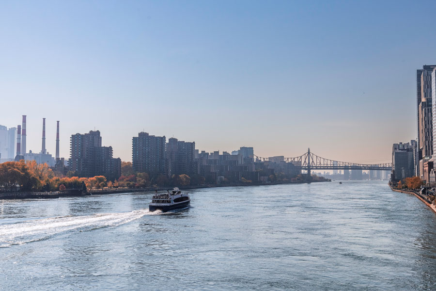 A passenger ferry travels north along the East River toward Roosevelt Island and the Queensboro Bridge on a clear autumn day in New York City. The scene captures the active waterway between Manhattan and Queens, with waterfront buildings, fall foliage, and the bridge forming a distinctive urban backdrop.