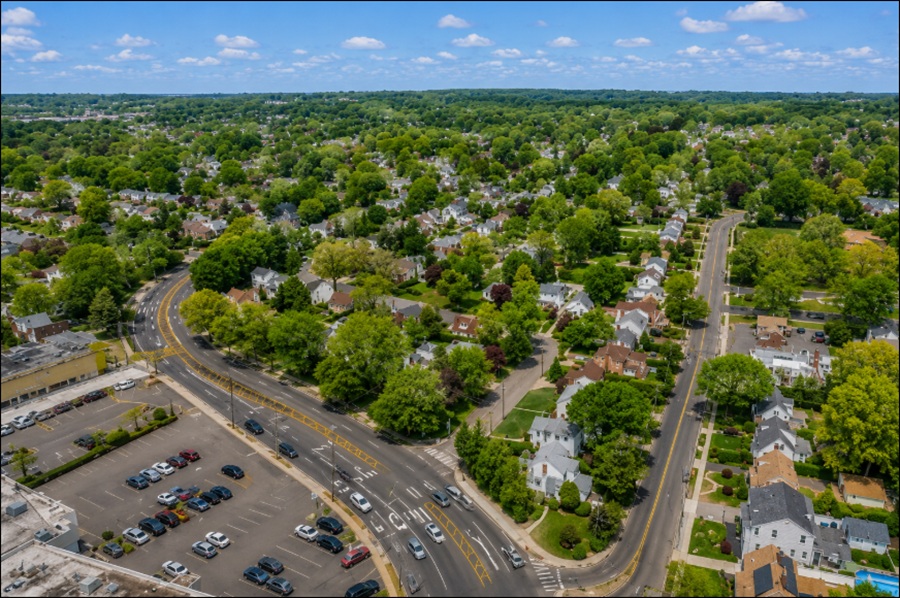 An aerial view of Manhasset Hills, New York, showcases a well-established suburban neighborhood with tree-lined streets, well-maintained single-family homes, and landscaped residential blocks. A major roadway borders the community alongside nearby commercial businesses and parking areas, illustrating the hamlet's convenient location and easy access to shopping, transportation, and other amenities in central Nassau County on Long Island.