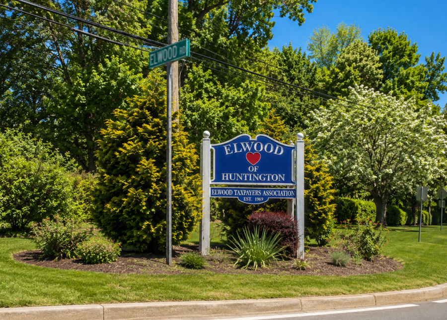 The Elwood of Huntington community sign welcomes visitors to Elwood, New York, a hamlet in the Town of Huntington on Long Island's North Shore. Surrounded by mature trees and landscaped greenery on a bright summer day, the entrance reflects the suburban character and well-established residential neighborhoods that define the community.