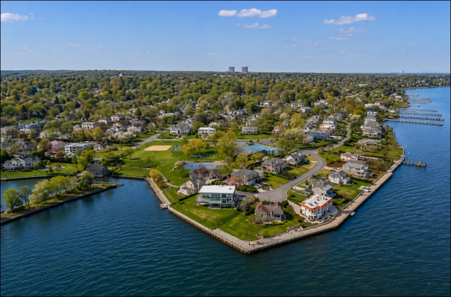 A sweeping aerial view showcases Harbor Hills, New York, with elegant waterfront homes, tree-lined residential streets, and manicured lawns overlooking the calm waters of the North Shore. Private docks, neighborhood parks, and a scenic shoreline highlight the community's coastal setting, while the surrounding landscape stretches inland toward dense woodlands and neighboring Nassau County communities.