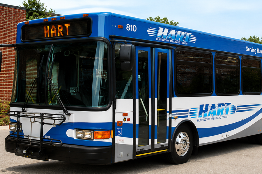 A modern Huntington Area Rapid Transit (HART) bus sits parked on a sunny day outside a brick building. The full-size low-floor transit bus features HART's distinctive blue-and-white livery with sweeping blue stripes, large HART logos along the side, and an amber LED destination sign reading "HART." Viewed from a front three-quarter angle, the bus showcases its large windshield, front bike rack, wide passenger doors, tinted windows, and accessible boarding design, highlighting the public transportation service that operates throughout the Town of Huntington on Long Island.