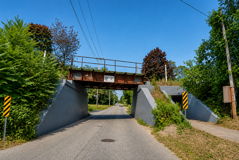 A Long Island Rail Road bridge carries the Main Line over a quiet roadway in Jamesport, reflecting the rural character of the North Fork. The low-clearance underpass is one of several railroad crossings that have served the community for generations.