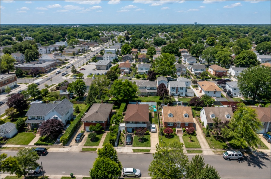 An aerial view of New Cassel, New York, overlooks a well-established residential neighborhood with a mix of single-family homes, tree-lined streets, and neatly maintained properties. Local businesses, neighborhood roads, and a nearby commercial corridor blend into the suburban landscape, illustrating the community's central Nassau County location and convenient access to shopping, transportation, and surrounding Long Island communities.