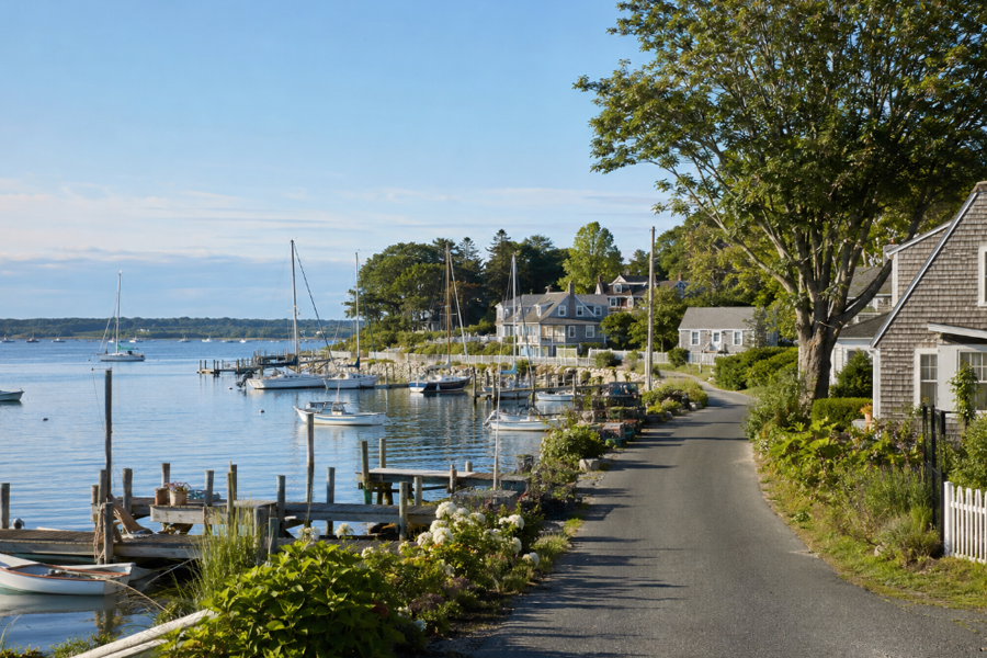 A quiet waterfront scene representative of New Suffolk, New York, with sailboats, private docks, coastal homes, and a narrow road along the harbor. The peaceful setting reflects the maritime character of this small North Fork community.
