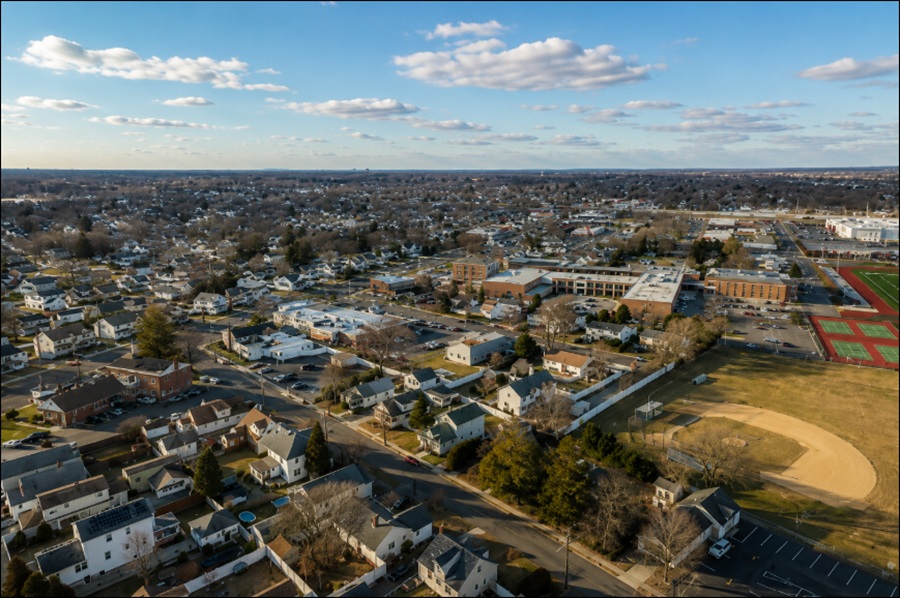 An aerial view of North Lindenhurst, New York, showcases a well-established suburban neighborhood with rows of single-family homes, local businesses, and community facilities surrounded by tree-lined streets. Nearby athletic fields, tennis courts, and commercial areas highlight the hamlet's family-friendly character and convenient location in western Suffolk County on Long Island.