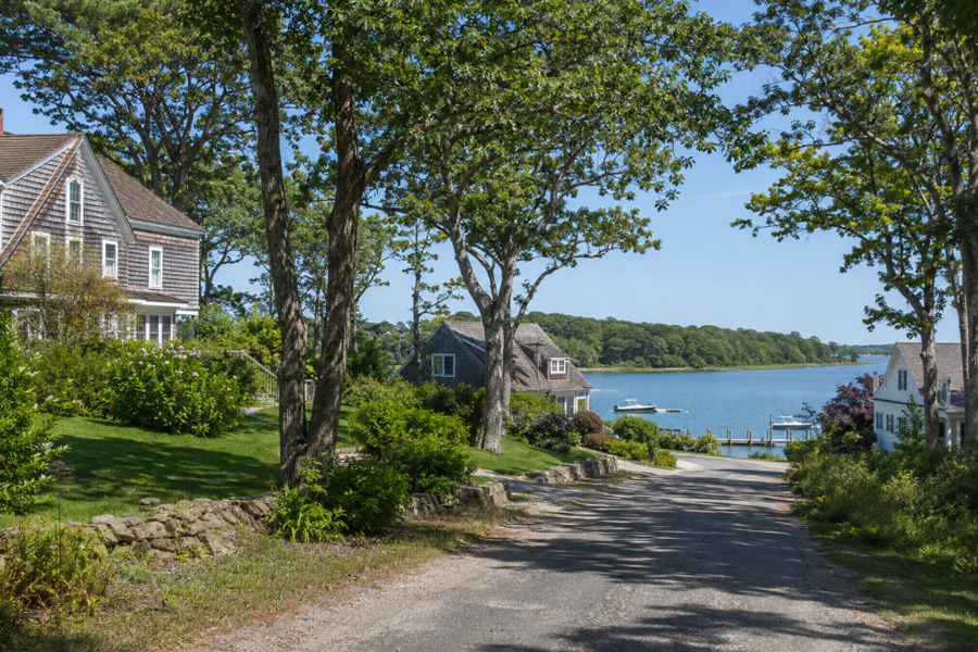 A tree-shaded residential road leads toward the calm waters of North Sea Harbor in North Sea, New York. Traditional shingled homes, mature landscaping, private docks, and small boats reflect the peaceful waterfront character of this Southampton community.