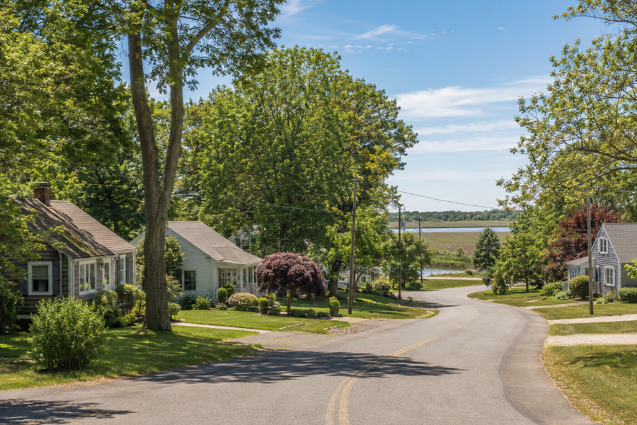 A quiet residential street in Riverside, New York, features modest homes surrounded by mature trees and natural greenery. The fictional scene reflects the community’s wooded East End setting and its close connection to the nearby Peconic River.