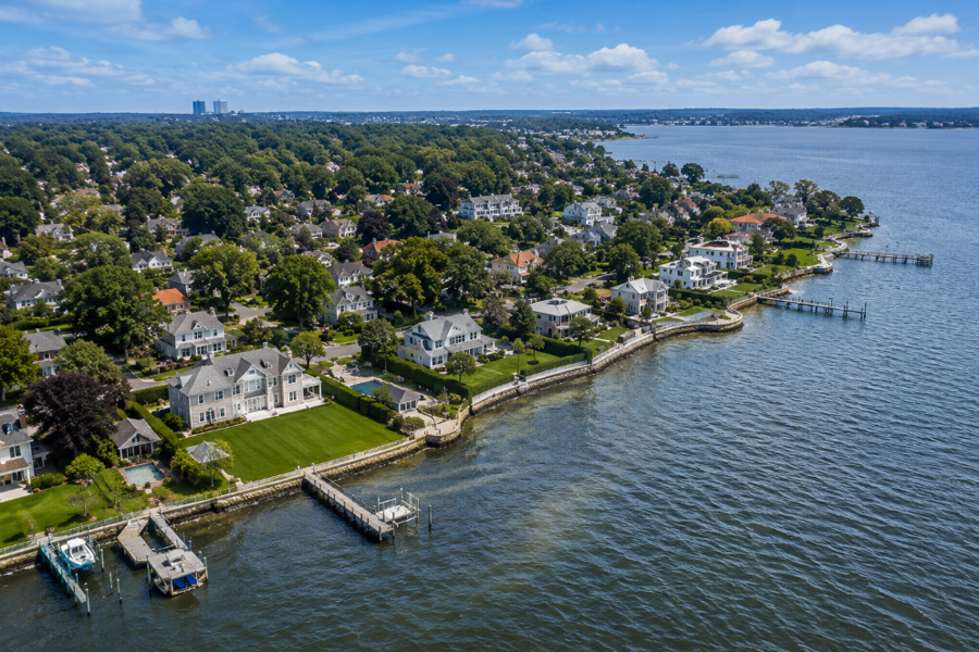 An aerial view of Saddle Rock showcases elegant waterfront homes along the shoreline of Little Neck Bay, where private docks, tree-lined streets, and manicured properties reflect the village's quiet residential character on the Great Neck Peninsula.