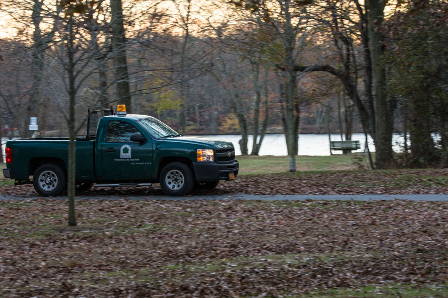 A New York State Parks maintenance vehicle travels through Hempstead Lake State Park, with one of the park's freshwater lakes and a quiet wooded landscape in the background. The park's extensive trail system, lakes, and natural areas are maintained year-round to support recreation, wildlife habitat, and public enjoyment.