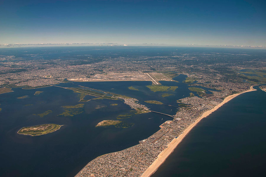 An aerial view of the Rockaway Peninsula in Queens, New York, separating Jamaica Bay (left) from the Atlantic Ocean (right). The image also shows John F. Kennedy International Airport along the northern shoreline of the bay, illustrating the unique mix of urban development, barrier beaches, wetlands, and open water that defines one of Long Island's most important coastal ecosystems.