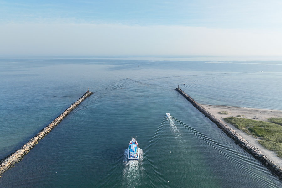 An aerial view of Montauk Inlet shows boats navigating between the stone jetties that connect Montauk Harbor with Block Island Sound and the Atlantic Ocean. The inlet is one of Long Island's busiest maritime gateways, serving commercial fishing vessels, charter boats, recreational boaters, and the United States Coast Guard throughout the year.