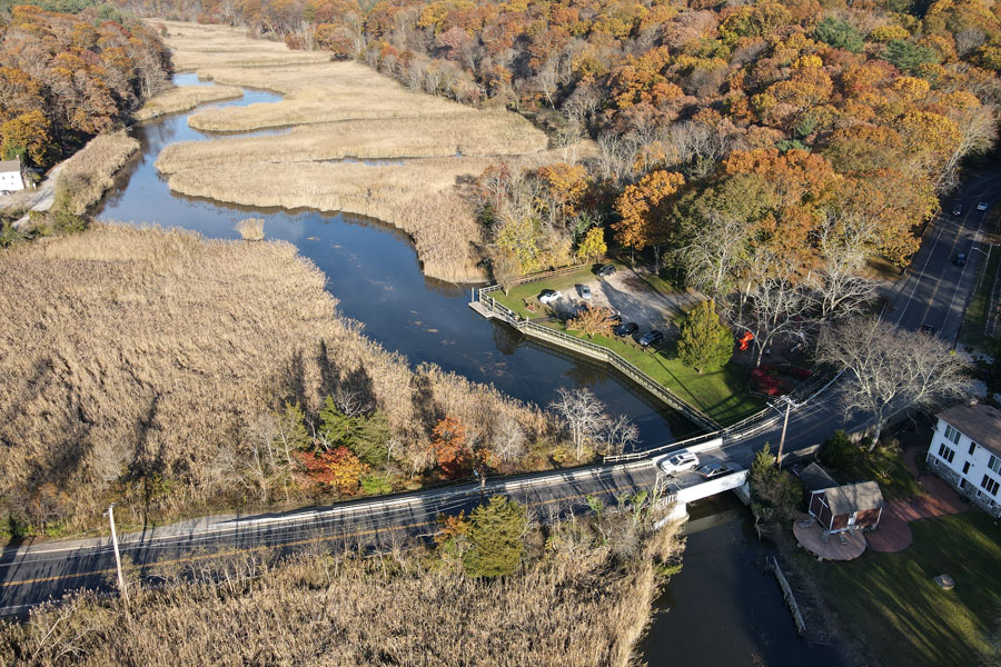 The Nissequogue River winds through expansive tidal marshes beneath a canopy of brilliant autumn foliage in the Village of Nissequogue. In the foreground, a small bridge carries local traffic across the river, illustrating the balance between peaceful residential life and one of Long Island's most scenic natural landscapes.