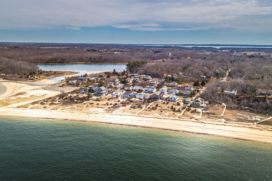This aerial view showcases a quiet stretch of Long Island's North Shore, where a sandy beach, protected cove, and waterfront homes border the calm waters of Long Island Sound. Surrounded by mature woodlands and natural shoreline, the community reflects the scenic coastal character that distinguishes the North Shore from the island's Atlantic-facing South Shore. The region is known for its sheltered harbors, picturesque waterfront villages, and peaceful landscapes that have attracted residents and visitors for generations.