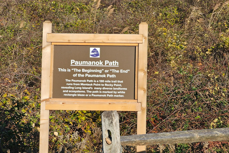 A wooden trailhead sign marks the beginning (or end) of the Paumanok Path, Long Island's renowned long-distance hiking trail. Surrounded by dense shrubs and native vegetation, the sign explains that the trail stretches approximately 130 miles between Montauk Point and Rocky Point, traversing many of Long Island's diverse landscapes and ecosystems. A rustic split-rail fence in the foreground reinforces the natural setting, welcoming hikers to one of the island's premier outdoor adventures. File photo: Patrick Hatt, licensed.