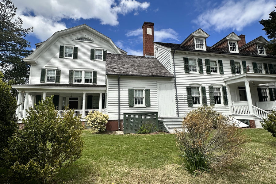 Sagtikos Manor displays its layered architectural history through a long white clapboard exterior, green shutters, brick chimneys, dormer windows, and multiple connected sections added over generations. Surrounded by lawn, shrubs, and mature trees, the preserved West Bay Shore estate offers a clear view of the craftsmanship and changing design styles that have shaped the historic home for more than three centuries.