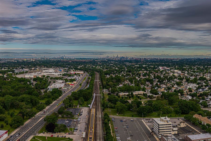 A sweeping aerial view looks west across a Long Island community toward the distant New York City skyline. The scene highlights the island's mix of residential neighborhoods, transportation corridors, green spaces, and its close connection to the city, making it a fitting overview for exploring Long Island's many sightseeing destinations.