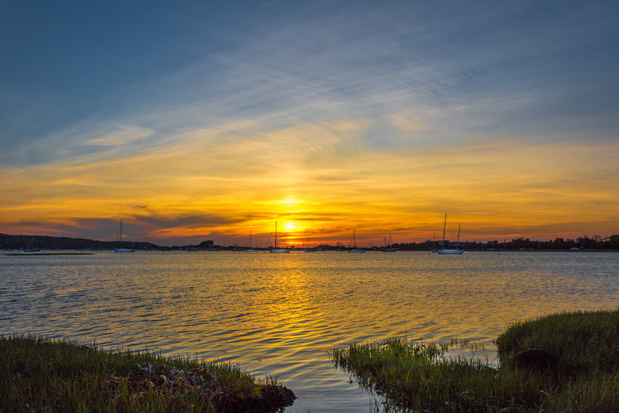 Sunset over Mount Sinai Harbor in Mount Sinai, New York, where anchored sailboats are silhouetted against a brilliant golden sky. The fading sunlight reflects across the calm harbor water, highlighting the peaceful character of this North Shore waterfront.