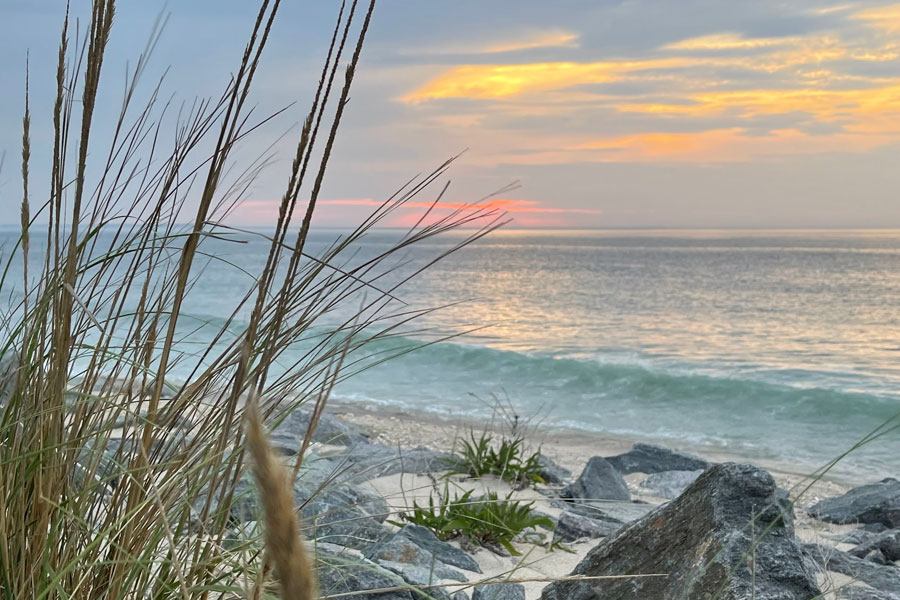 Sea oats sway above the rocky shoreline as gentle waves roll onto the beach at sunset along the Long Island Sound in Sound Beach. The hamlet's scenic waterfront is known for its coastal bluffs, peaceful shoreline, and sweeping views across the Sound toward the Connecticut coast.