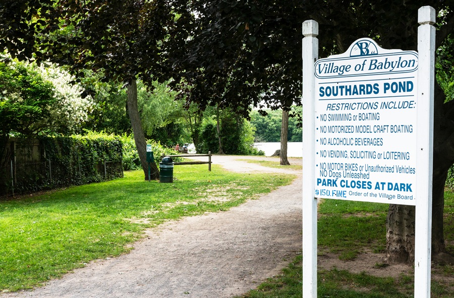An entrance sign welcomes visitors to Southards Pond in the Village of Babylon, where a tree-lined walking path leads toward the scenic freshwater pond. The park is a popular destination for walking, fishing, birdwatching, and enjoying the peaceful natural setting, while posted regulations help protect the area's wildlife and preserve its quiet recreational atmosphere. File photo: WoodysPhotos, licensed.