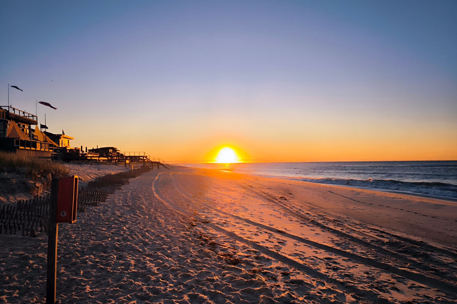 A brilliant sunrise breaks over the Atlantic Ocean at Fire Island, casting warm orange and gold light across the beach while the surrounding sky remains a cool shade of blue. The rising sun reflects across the water and wet sand, creating a vivid contrast between the glowing horizon and the quiet early-morning shoreline. File photo: Diane BerlinX7, licensed.